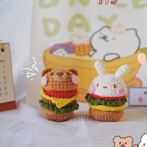 以下が含まれることがあります： 手作りの編みぐるみハンバーガーのおもちゃが2つ。1つはクマの顔、もう1つはウサギの顔。ハンバーガーは茶色、赤、黄色、緑の層になっています。背景にはカレンダーと漫画のイラストがあります。