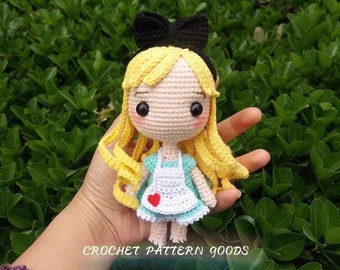 May include: A crocheted doll of Alice from Alice in Wonderland. She has blonde hair, a blue dress, a white apron with a red heart, and a black bow. The doll is being held in a hand against a green background.