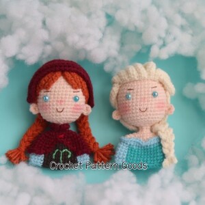 May include: Two crocheted dolls, one with red hair and a burgundy cape, the other with blonde hair and a blue dress. The dolls are sitting on a white fluffy surface with the text "Crochet Pattern Goods" below them.