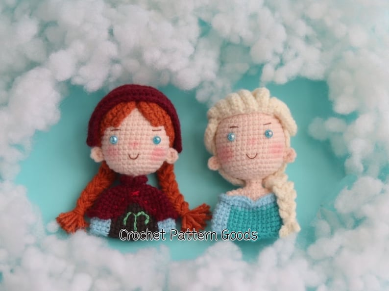 May include: Two crocheted dolls, one with red hair and a burgundy cape, the other with blonde hair and a blue dress. The dolls are sitting on a white fluffy surface with the text "Crochet Pattern Goods" below them.