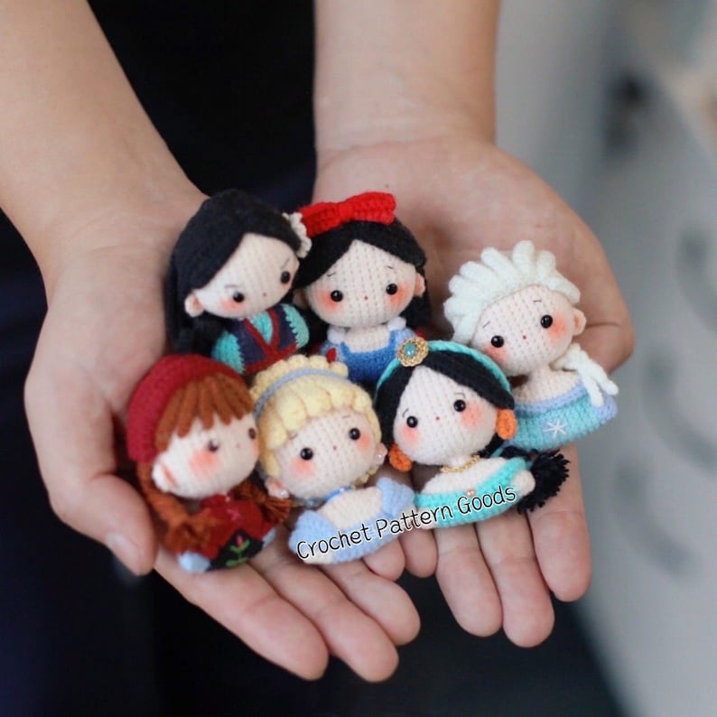 May include: A collection of seven crocheted Disney princess dolls. The dolls are all wearing their signature dresses and have detailed facial features. The dolls are arranged in a circle, with the text "Crochet Pattern Goods" visible in the bottom center of the image.