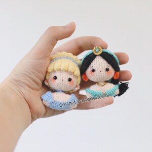 May include: Two crocheted dolls, one with blonde hair and a blue dress, the other with black hair and a teal dress. Both dolls have embroidered faces and are wearing gold jewelry.