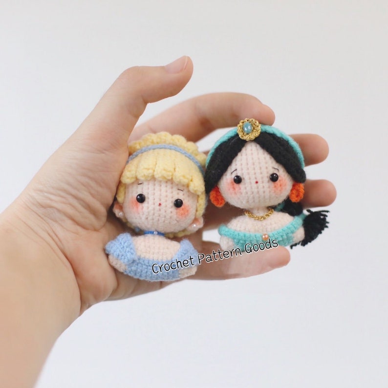 May include: Two crocheted dolls, one with blonde hair and a blue dress, the other with black hair and a teal dress. Both dolls have embroidered faces and are wearing gold jewelry.