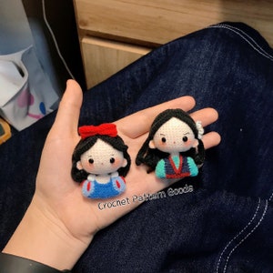 May include: Two crocheted dolls, one with a red bow and blue dress, the other with a green and blue dress. The dolls are on a hand with the text "Crochet Pattern Goods" below them.