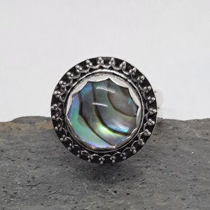 May include: A silver ring with a round abalone shell inlay. The ring has a decorative border around the shell.