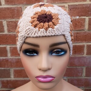 May include: A beige crochet hat with a sunflower design in shades of brown. The hat is made of a textured yarn and features a central flower with a dark brown centre and lighter brown petals. The hat is displayed on a mannequin.