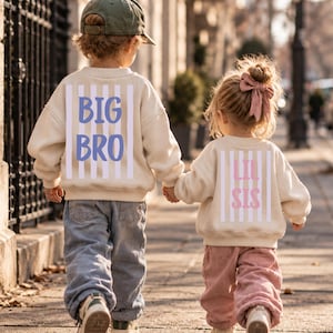 Könnte beinhalten: Zwei cremefarbene Sweatshirts mit vertikalen weißen Streifen und Text auf der Rückseite. Ein Sweatshirt zeigt den blauen Text BIG BRO, das andere den rosa Text LIL SIS. Beide werden mit locker sitzenden Hosen und Turnschuhen getragen.