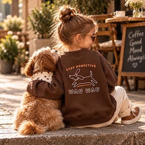 Könnte beinhalten: Ein dunkelbraunes Langarm-Sweatshirt mit einer Hunde-Grafik und dem Text "STAY PAWSITIVE" sowie "WAU WAU" auf der Rückseite. Das Sweatshirt hat einen Rundhalsausschnitt und gerippte Bündchen, kombiniert mit einer hellen Hose und braunen Sneakern.