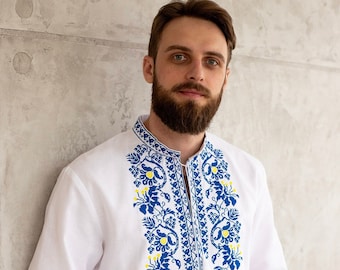 Könnte beinhalten: Ein weißes Leinenhemd mit blauen und gelben gestickten Blumenmustern. Das Hemd hat lange Ärmel und einen Knopfkrag.