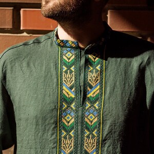 K&ouml;nnte beinhalten: Ein dunkelgr&uuml;nes Leinenhemd mit einem traditionellen Stickmuster in Gelb, Blau und Gr&uuml;n. Die Stickerei zeigt geometrische Formen und stilisierte Blumenmotive.