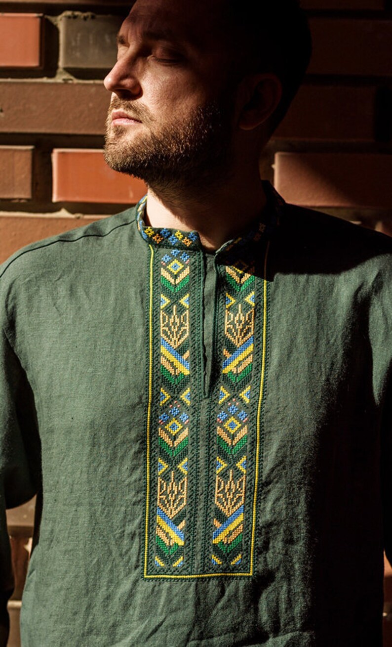 K&ouml;nnte beinhalten: Ein dunkelgr&uuml;nes Leinenhemd mit einem traditionellen Stickmuster in Gelb, Blau und Gr&uuml;n. Die Stickerei zeigt geometrische Formen und stilisierte Blumenmotive.