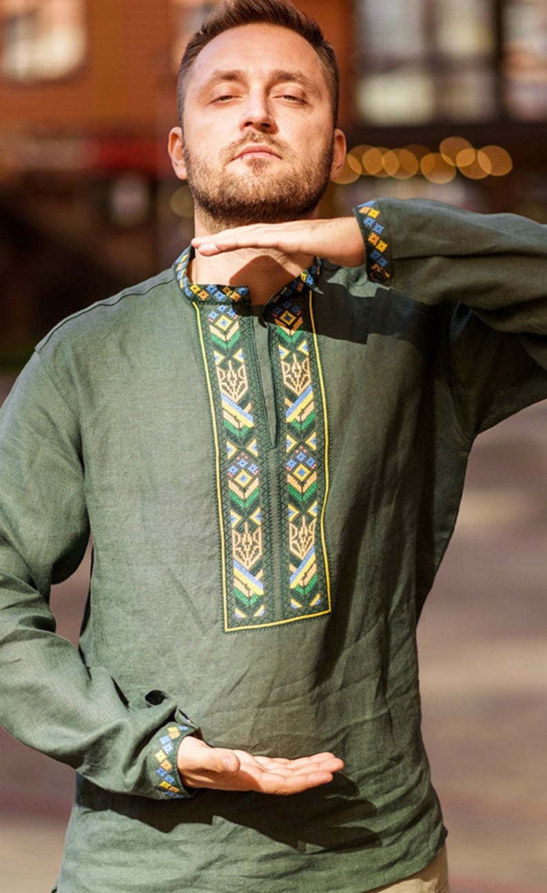 K&ouml;nnte beinhalten: Ein Mann tr&auml;gt ein gr&uuml;nes Leinenhemd mit Stickereien. Die Stickerei zeigt ein traditionelles ukrainisches Muster in Gelb, Blau und Gr&uuml;n.