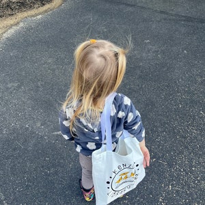 May include: A young child wearing a grey and white patterned sweater and striped leggings carries a white tote bag with the words "Kenzi's Library" and a colorful illustration of a book and birds.