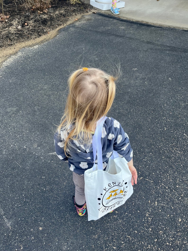 May include: A young child wearing a grey and white patterned sweater and striped leggings carries a white tote bag with the words "Kenzi's Library" and a colorful illustration of a book and birds.