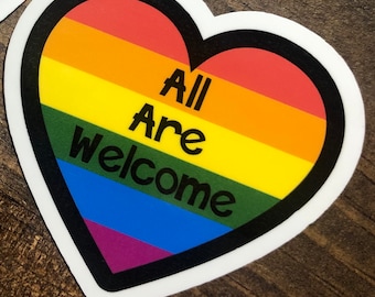 Può includere: Un adesivo a forma di cuore arcobaleno con un contorno nero. L'adesivo dice "All Are Welcome" in testo nero.