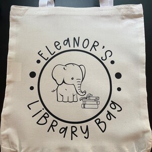 May include: White canvas tote bag with black text and an illustration of an elephant holding a stack of books. The text reads "Eleanor's Library Bag."