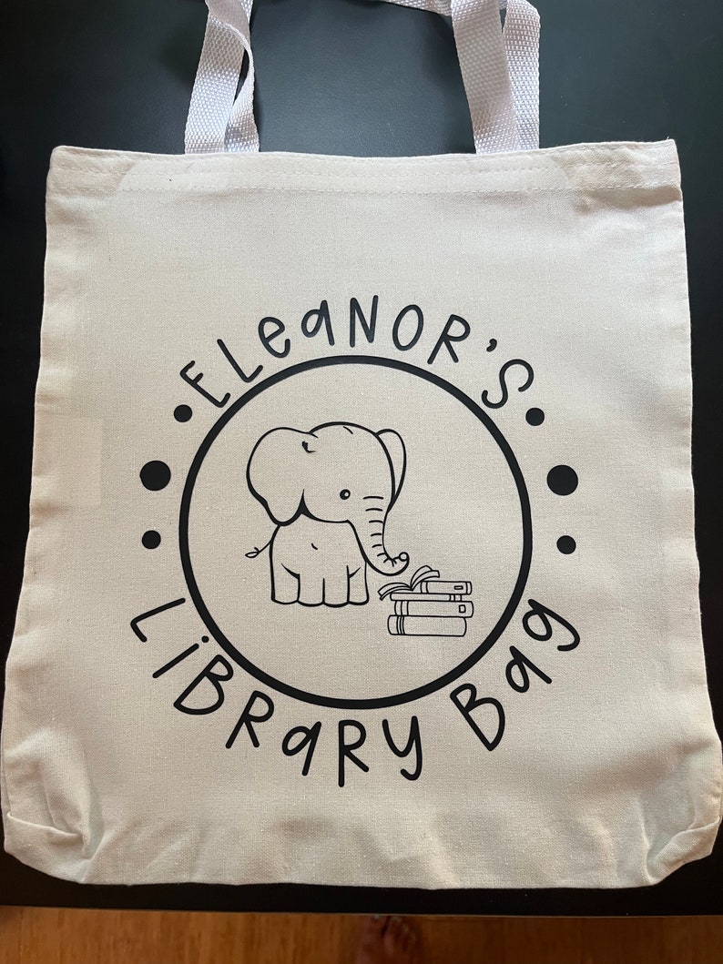 May include: White canvas tote bag with black text and an illustration of an elephant holding a stack of books. The text reads "Eleanor's Library Bag."