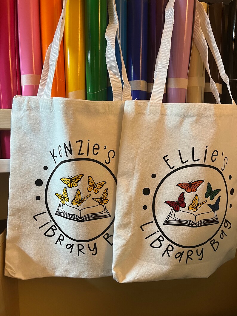 May include: Two white canvas tote bags with black handles. The bags have a black circle design with a book and butterflies. One bag says "Kenzie's Library Bag" and the other says "Ellie's Library Bag".