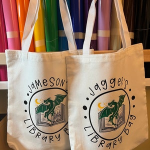 May include: Two white canvas tote bags with black and green dragon designs. The left bag says "Jameson's Library Bag" and the right bag says "Jagger's Library Bag".