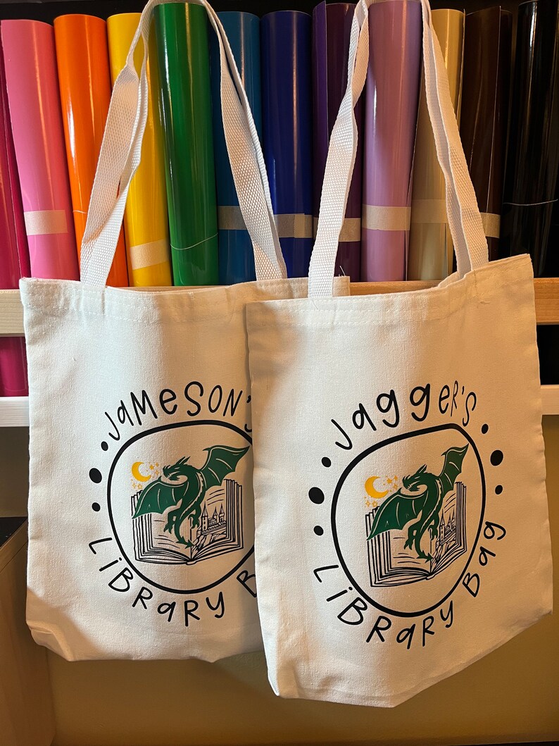 May include: Two white canvas tote bags with black and green dragon designs. The left bag says "Jameson's Library Bag" and the right bag says "Jagger's Library Bag".