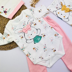 Może przedstawiać: Biały śpioszek dla niemowlęcia z nadrukiem różowego króliczka i wzorem kwiatowym. Śpioszek jest połączony z różowymi spodniami.