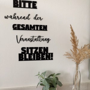 May include: Black vinyl lettering on a white wall reads "Bitte" (Please), "w&auml;hrend der" (during the), "GESAMTEN" (ENTIRE), "Veranstaltung" (Event), "SITZEN" (SIT), "BLEIBEN!" (STAY!).
