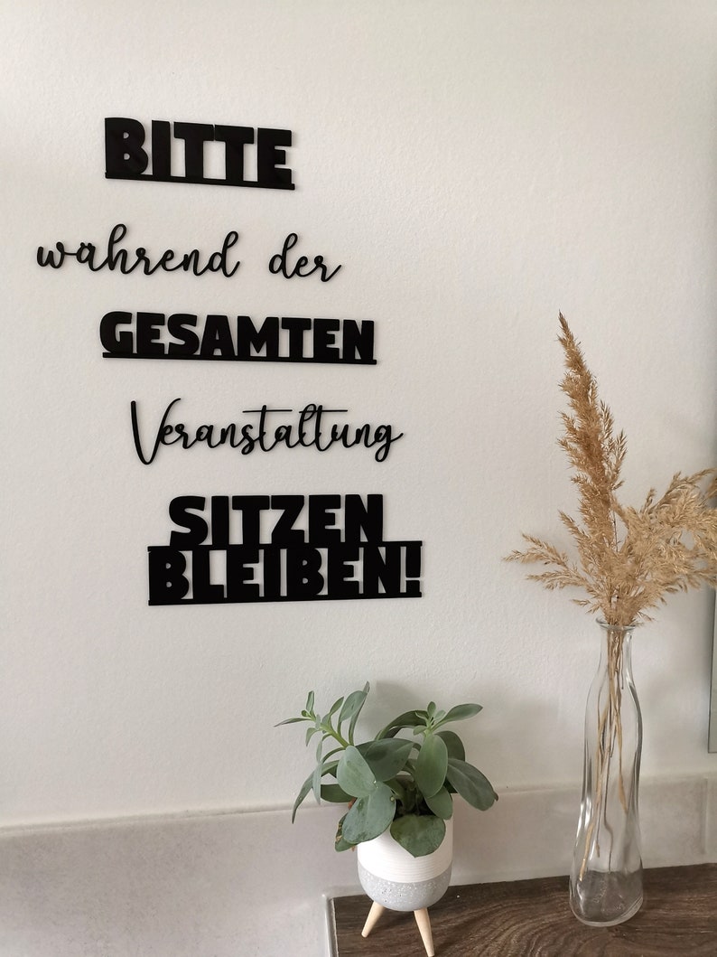 May include: Black vinyl lettering on a white wall reads "Bitte" (Please), "w&auml;hrend der" (during the), "GESAMTEN" (ENTIRE), "Veranstaltung" (Event), "SITZEN" (SIT), "BLEIBEN!" (STAY!).