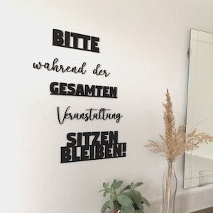 May include: Black and white sign with the text "Bitte w&auml;hrend der gesamten Veranstaltung sitzen bleiben!" on a white wall.