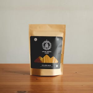 Pode incluir: Um saco de papel castanho de cacau Golden Milk. O saco tem um rótulo preto com as palavras "MAYA MOON Cacao" e "GOLDEN MILK". O saco está sobre uma superfície de madeira.