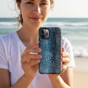 Puede incluir: Funda de teléfono azul oscuro con un patrón de puntos blancos, sostenida por una persona. La funda tiene un acabado brillante y un recorte para la cámara. El fondo muestra una playa y el océano.
