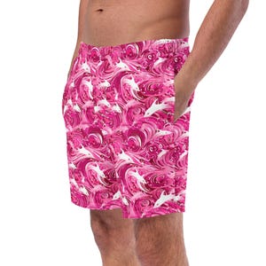 May include: Pink swim trunks with a repeating pattern of white dolphins swimming in pink waves. The shorts have a drawstring waist and side pockets. The fabric is a vibrant pink with darker pink and white accents.