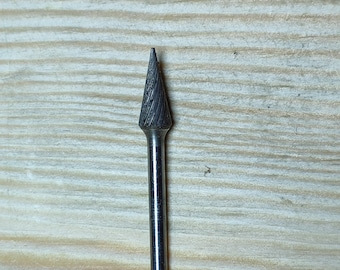 May include: A silver-colored rotary burr with a conical head and a cylindrical shank. The head has a spiral-grooved texture. The tool is designed for shaping and smoothing materials. The burr is shown against a light brown wooden surface.