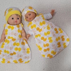 May include: Two baby dolls wearing matching yellow and white sleep sacks with a chick pattern. The dolls are wearing yellow knitted hats.