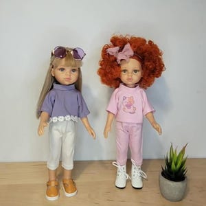 May include: Two dolls are displayed, each dressed in stylish outfits. One doll wears a lavender top, white pants, and brown shoes, with sunglasses. The other doll is in a pink outfit and white boots. A small potted plant is also present.