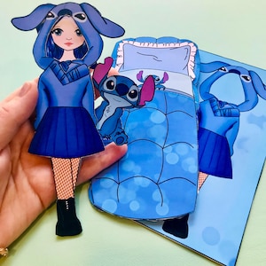 May include: A blue paper doll featuring a Stitch-themed outfit, including a hooded sweatshirt, skirt, and boots. A matching bed and Stitch character are also present. The doll is held in a hand.