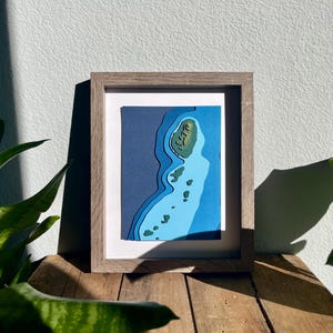 Puede incluir: Una pieza de arte en papel enmarcada que representa una isla azul y verde con un efecto 3D. La isla está rodeada de agua y tiene una pequeña colina verde en el centro.