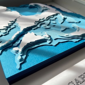 May include: A blue and white paper art piece depicting a coastal landscape. The piece is layered to create a 3D effect, with the white paper representing land and the blue paper representing water. The text "CAPE COD" is visible at the bottom of the image.