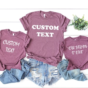 Pode incluir: Tr&ecirc;s camisetas marrons com o texto "CUSTOM TEXT" em branco. As camisetas est&atilde;o dispostas em fila, com a camiseta maior no meio e a menor &agrave; direita. As camisetas est&atilde;o sobre um fundo de madeira branco.