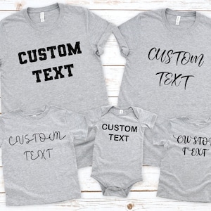 Pode incluir: Quatro t-shirts cinzas com o texto "CUSTOM TEXT" em preto. As t-shirts s&atilde;o de tamanhos e estilos diferentes, uma delas &eacute; um macac&atilde;o para beb&ecirc;.