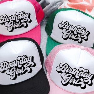 May include: Four trucker hats with a white front panel and a mesh back. The front panel of each hat has the text "Birthday Girl" in a retro, black font. The hats are in pink, black, green, and pink.