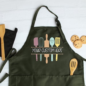 May include: A dark green apron with two large pockets and a design of kitchen utensils, including a whisk, rolling pin, spatula, and spoon. The text "YOUR CUSTOM HERE" is printed on the apron.