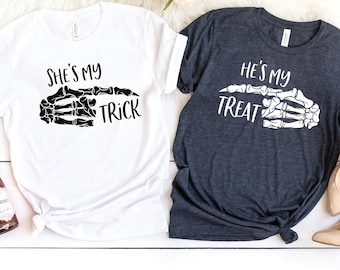 May include: Two t-shirts, one white and one dark gray, with Halloween-themed graphics. The white shirt says "She's my Trick" and the dark gray shirt says "He's my Treat", both with a skeleton hand design. The shirts are on a white surface.
