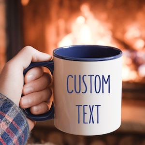 May include: A white ceramic mug with a blue interior and handle, featuring the words "CUSTOM TEXT" in blue. The mug is held by a person, with a cozy fireplace in the background.