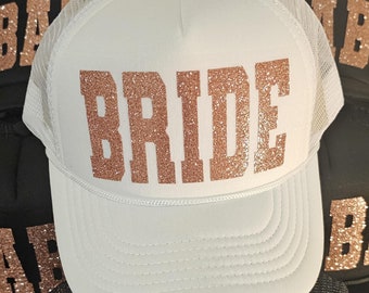 May include: White trucker hat with a mesh back and a curved brim. The word "BRIDE" is displayed in large, glittery, rose gold letters across the front. Other black hats with rose gold lettering are visible in the background.