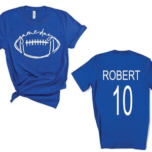 Op de afbeelding: Koningsblauw t-shirt met witte tekst en een football-afbeelding met de tekst "gameday". De achterkant van het shirt heeft de naam "ROBERT" en het nummer "10" in wit.