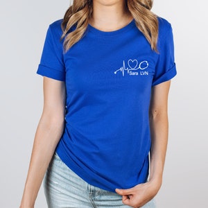 K&ouml;nnte beinhalten: Ein k&ouml;nigsblaues T-Shirt mit einer wei&szlig;en Grafik einer Herzschlaglinie, einem Stethoskop und dem Text "Sara LVN".