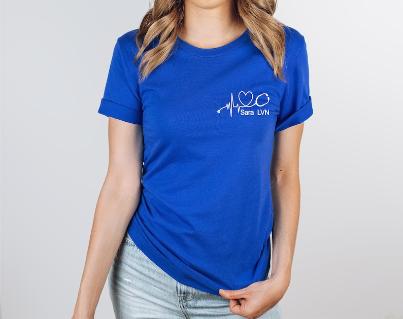 K&ouml;nnte beinhalten: Ein k&ouml;nigsblaues T-Shirt mit einer wei&szlig;en Grafik einer Herzschlaglinie, einem Stethoskop und dem Text "Sara LVN".