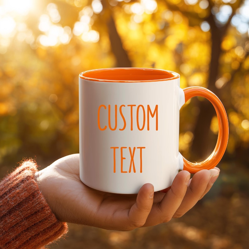 May include: White ceramic mug with an orange interior and handle, held in a hand. The mug features the words "CUSTOM TEXT" in orange lettering. The background is a blurred view of fall foliage.