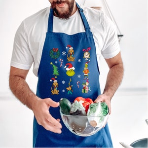Op de afbeelding: Een marineblauwe schort met kerstcartoonfiguren en symbolen. De schort heeft een feestelijk ontwerp met afbeeldingen van de Grinch, honden en andere vakantie-elementen. De schort wordt door een persoon vastgehouden.