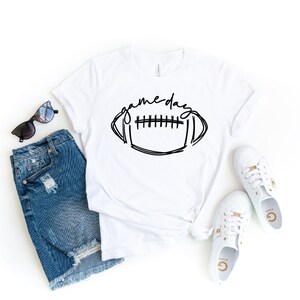 May include: White t-shirt featuring a black and white illustration of a football and the text "game day". The shirt is styled with denim shorts, sunglasses, and white trainers.